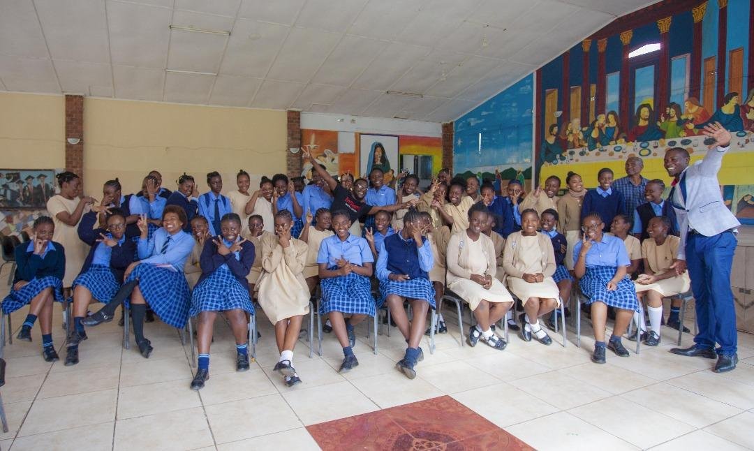 A large group of secondary school girls in blue and beige uniform seated together, laughing, with an MCUSzambia facilitator standing beside them